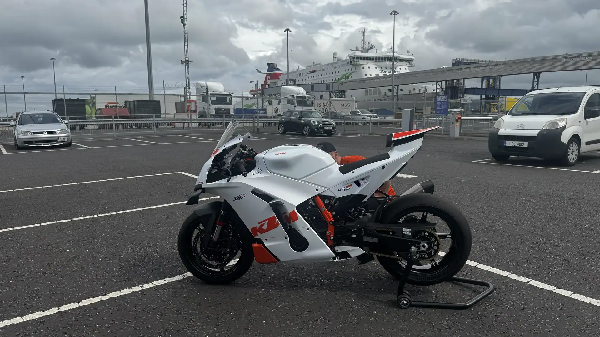 A white and orange KTM 990 RC R on a paddock stand in the port car park in Belfast, with a Stena Line ferry behind.