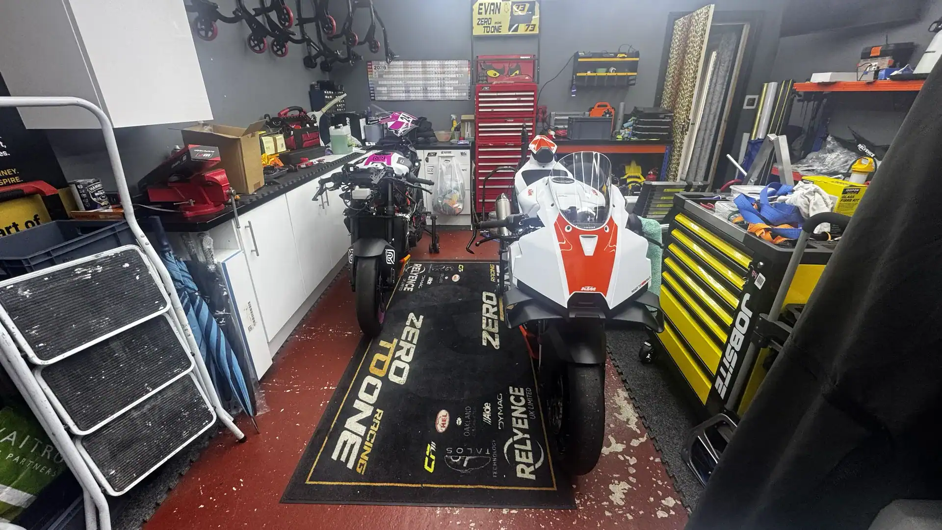 KTM 990 RC R Acquisition and New Livery 3 The white and orange KTM 990 RC R standing on a Zero To One Racing mat in the team garage, beside Tom's pink and white number 73 KTM 990 RC R.