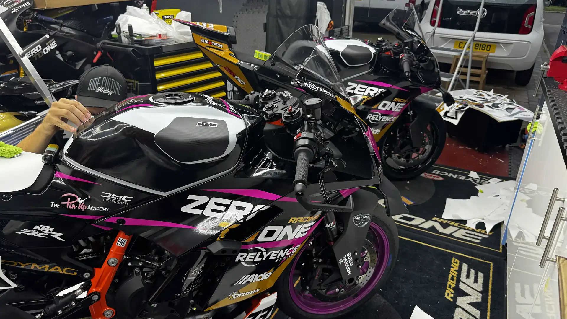 KTM 990 RC R Acquisition and New Livery 4 New pink, gold and black Zero To One Racing graphics being applied to a KTM 990 RC R in the garage, with a second 990 RC R behind and used decal backing sheets on the floor.